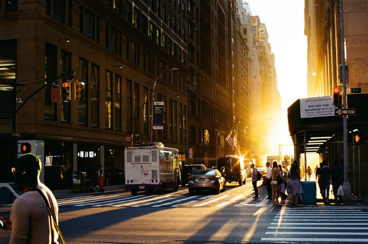 A street in New York is illuminated by the golden light of a sunset, with people waiting to cross and vehicles driving on the road.