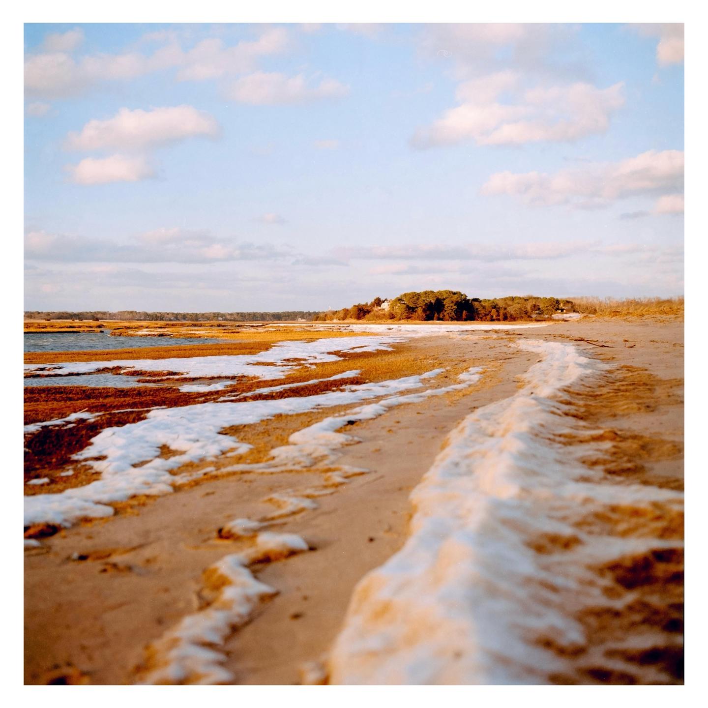 A sandy beach with patches of snow and marsh grass leads to a body of water under a cloudy sky.