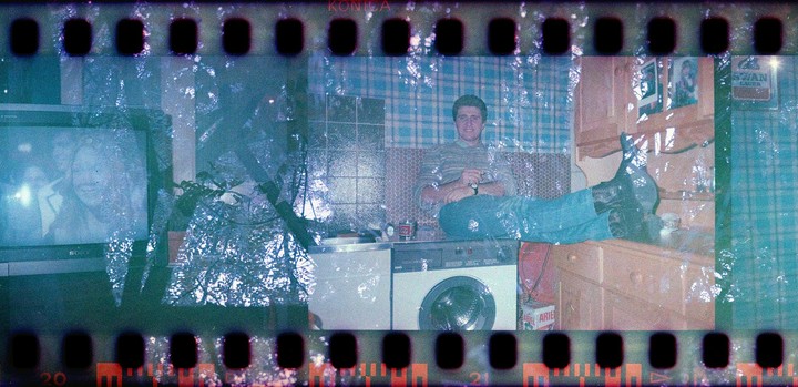 A man is sitting on a washing machine with his feet up on a wooden dresser, with a television and a plant visible in the room, and a double exposure of trees.
