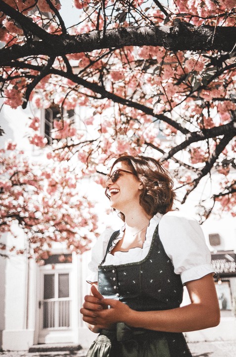A woman in a dirndl laughs under a cherry blossom tree.