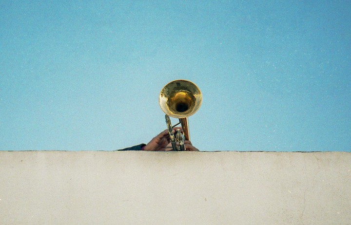 A person's hands hold a brass trumpet above a concrete wall against a clear blue sky.