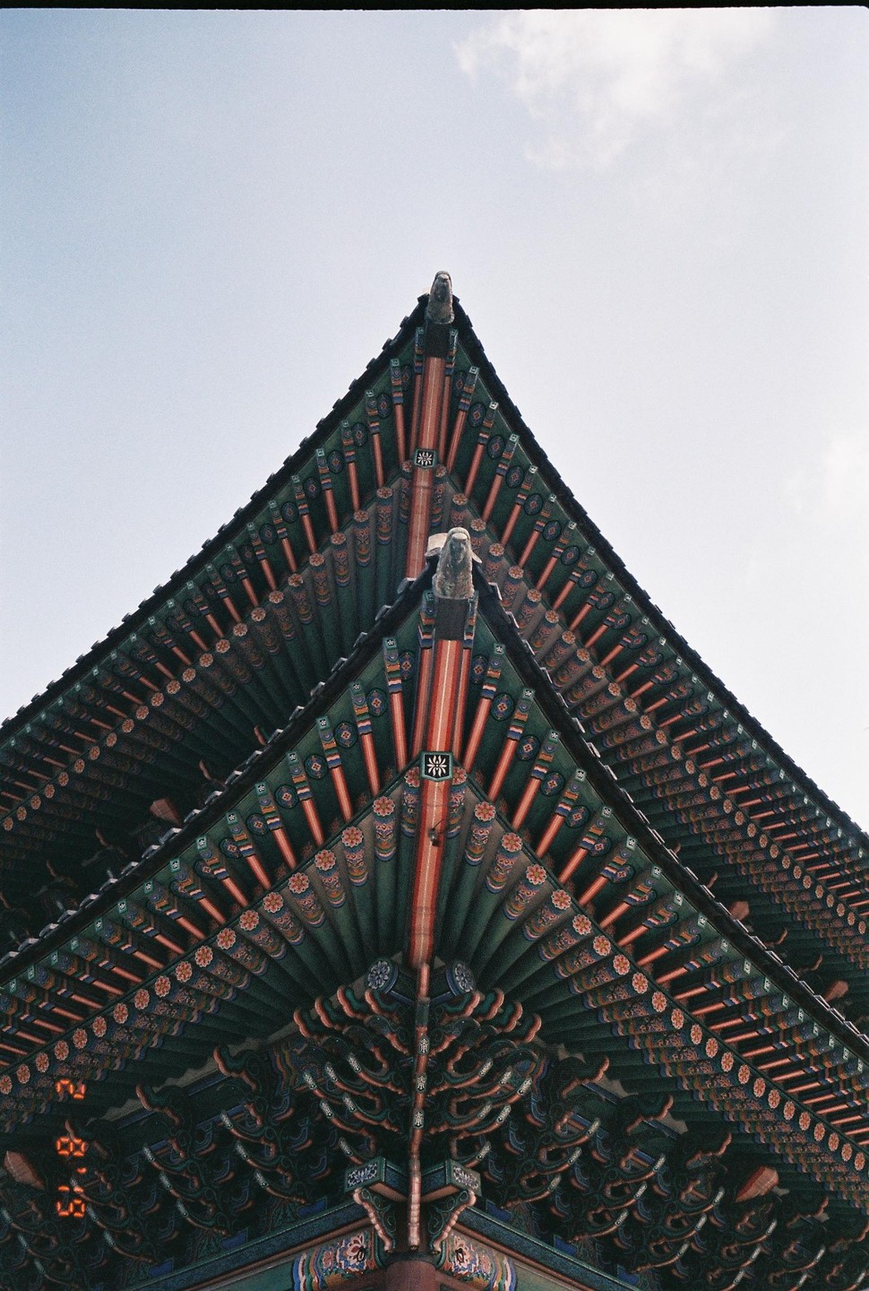 The ornate, colorful roof of a traditional Korean palace is viewed from below against a pale sky.