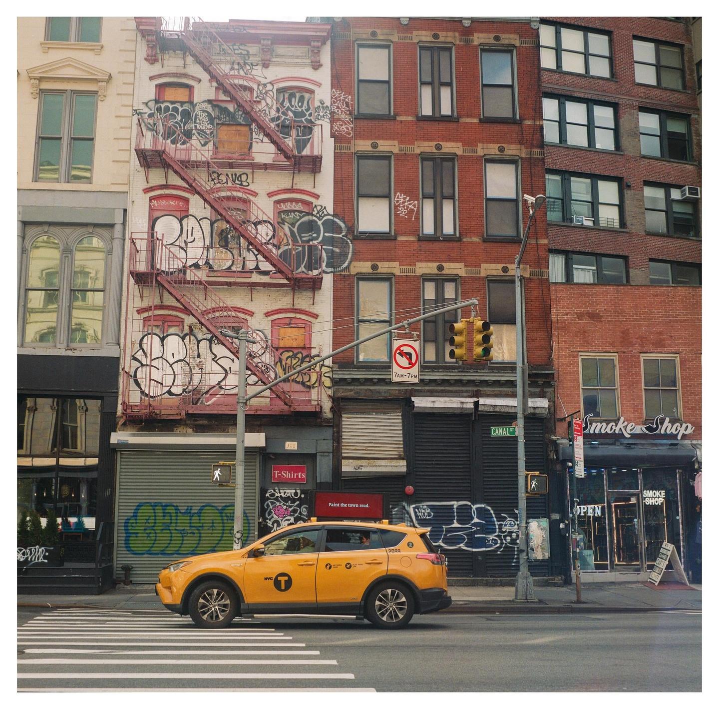 A yellow taxi drives across a crosswalk in front of graffiti-covered buildings with fire escapes.