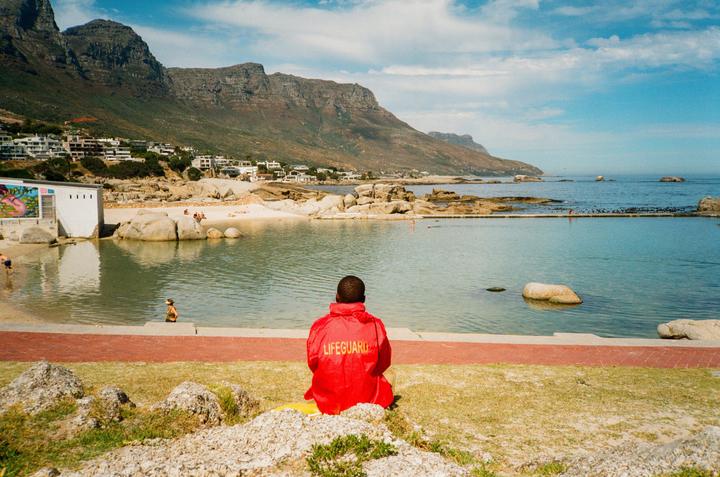 A lifeguard in a red jacket sits on a grassy bank overlooking a beach and ocean with mountains in the background.