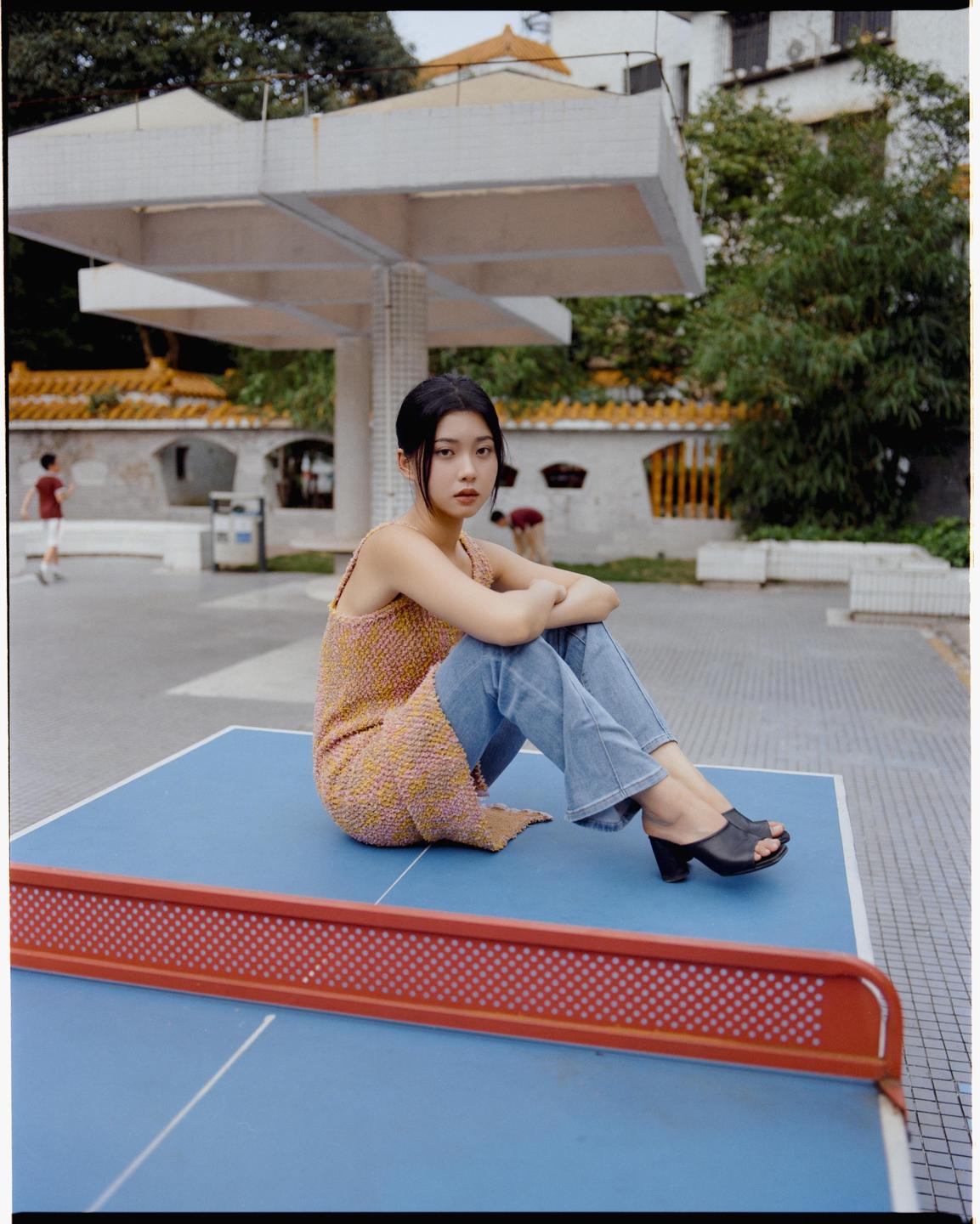 A woman sits on a blue ping-pong table with a red net, looking at the viewer.