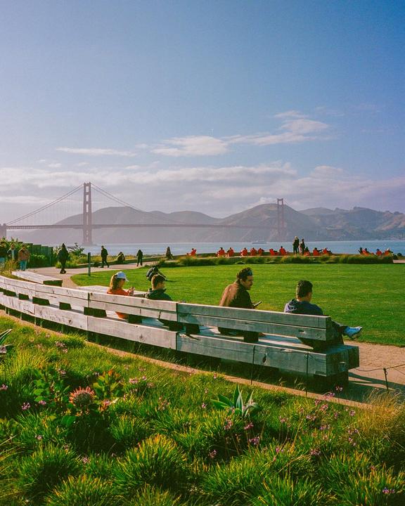 People sit on a long wooden bench and on a grassy hill overlooking the Golden Gate Bridge and bay.