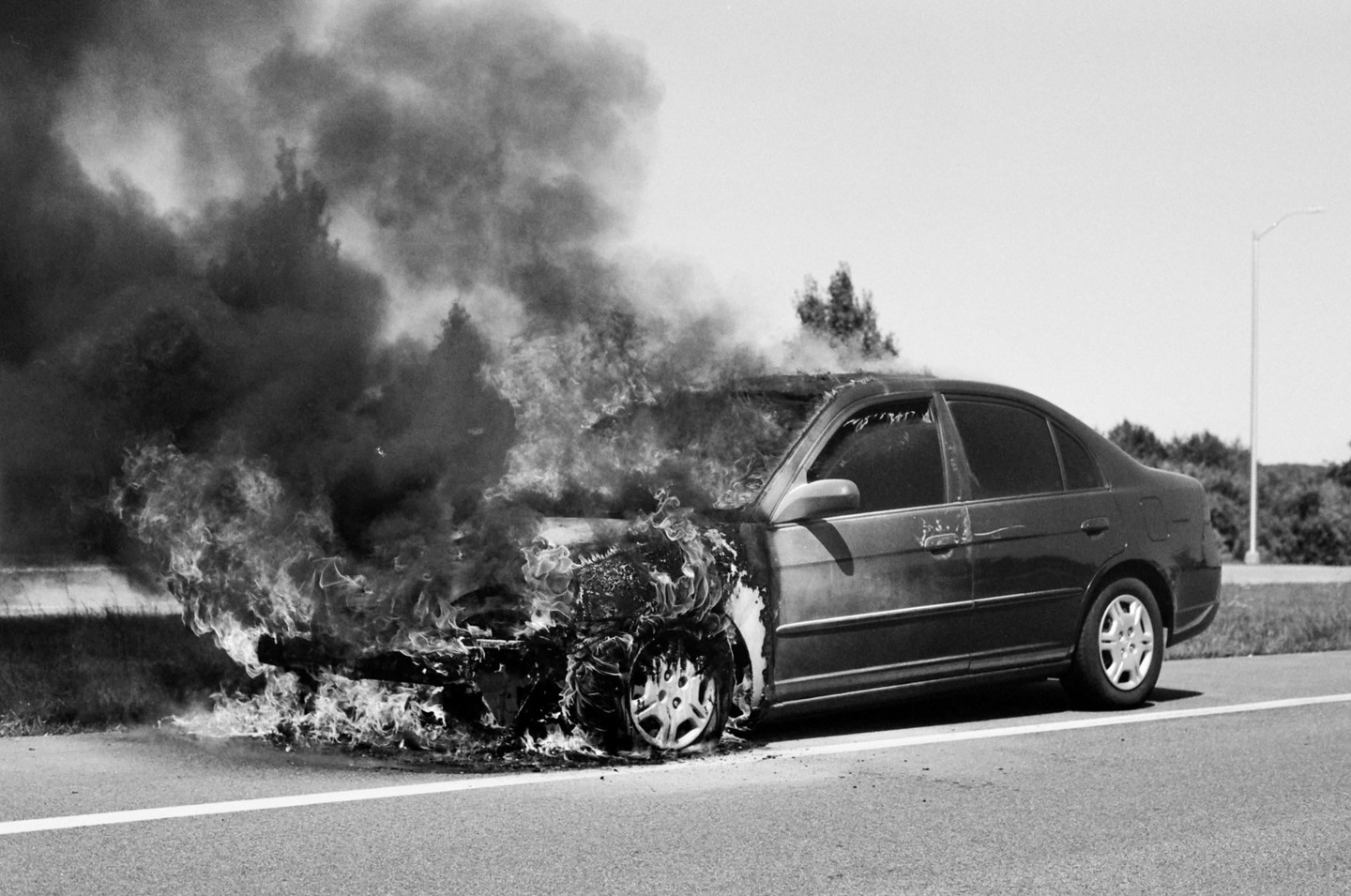 A car is engulfed in flames and thick smoke on the side of a road.