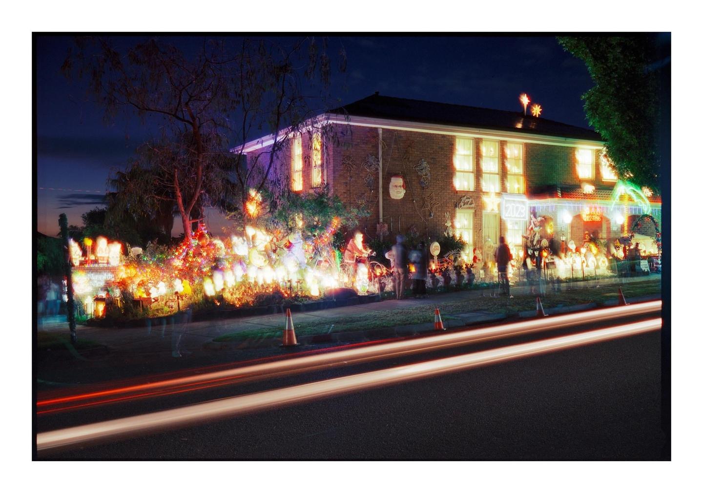 A house is decorated with many colorful Christmas lights and figures at night, with light trails from passing vehicles in the foreground.