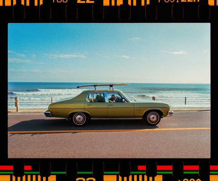 A green car with a surfboard on its roof drives along a coastal road next to the ocean.