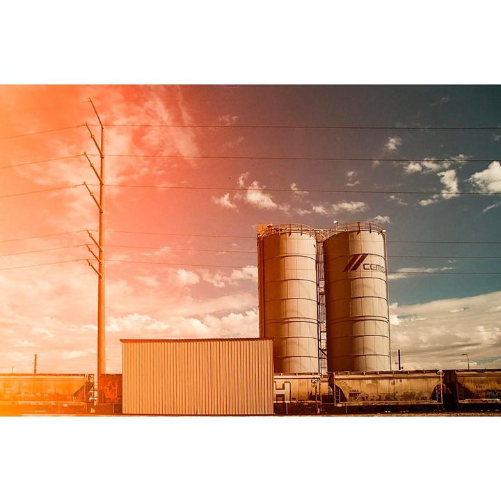 Two large industrial silos stand next to a train with power lines and a utility pole under a partly cloudy sky.
