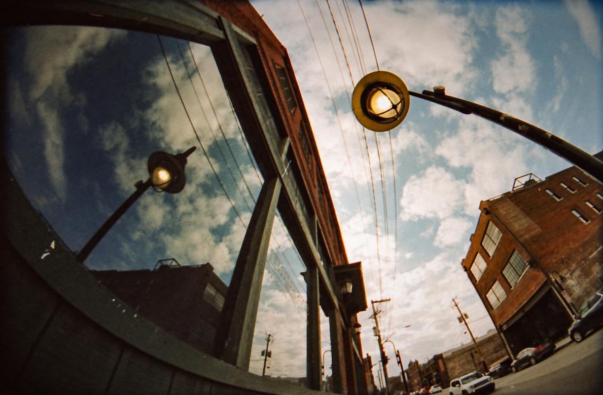 A street lamp and cloudy sky are reflected in a window, with a brick building and street visible in the background.