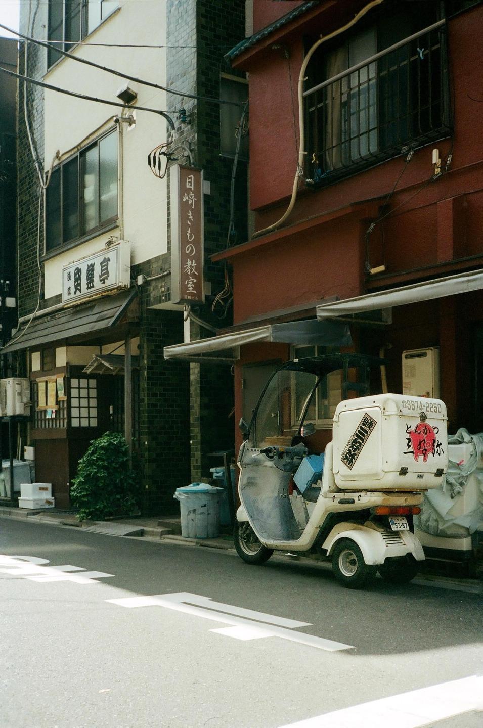 A white delivery scooter is parked on a street in front of buildings with Japanese signs.
