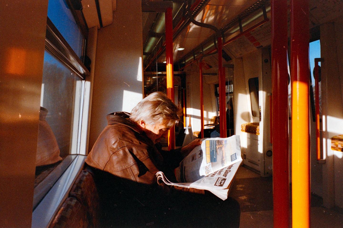 A man in a brown leather jacket reads a newspaper on a train with sunlight streaming through the windows.