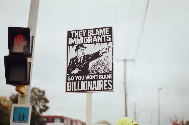 A protest sign with the text "They blame immigrants so you won't blame billionaires" is displayed next to a traffic light.