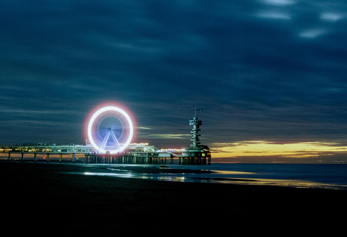 A ferris wheel with light trails and a pier are visible from a beach at dusk.