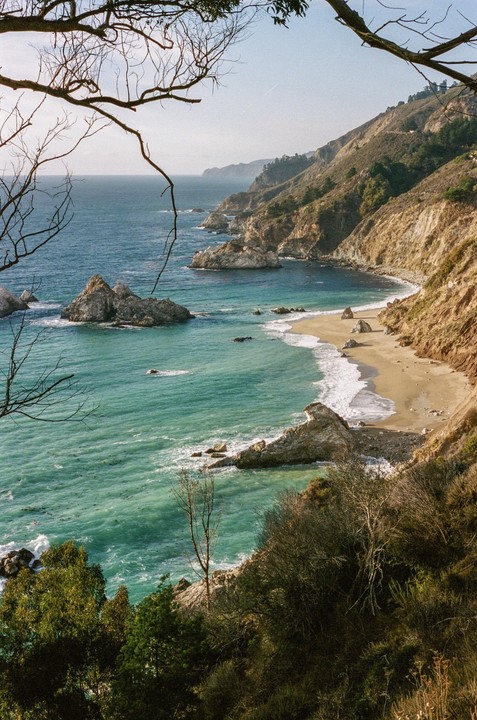 A scenic view of the Big Sur coastline shows a sandy beach, turquoise water, and rocky cliffs under a clear sky.