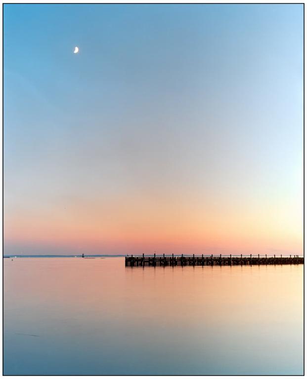 A crescent moon hangs in the sky above a pier stretching into calm water at dusk.