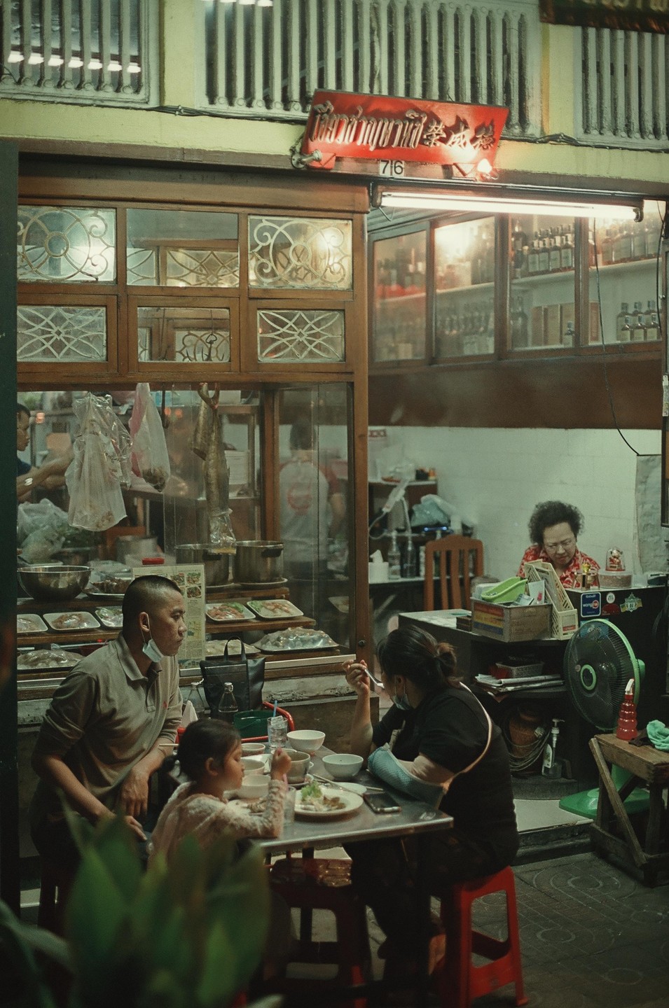 A family eats at a table outside a restaurant with a woman sitting inside.