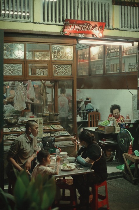 A family eats at a table outside a restaurant with a woman sitting inside.