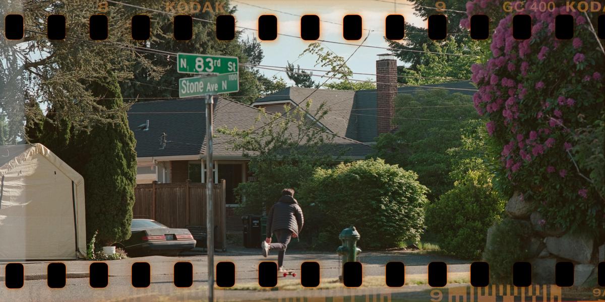 A person on a skateboard moves away from the camera on a residential street with a street sign for N. 83rd St and Stone Ave N.