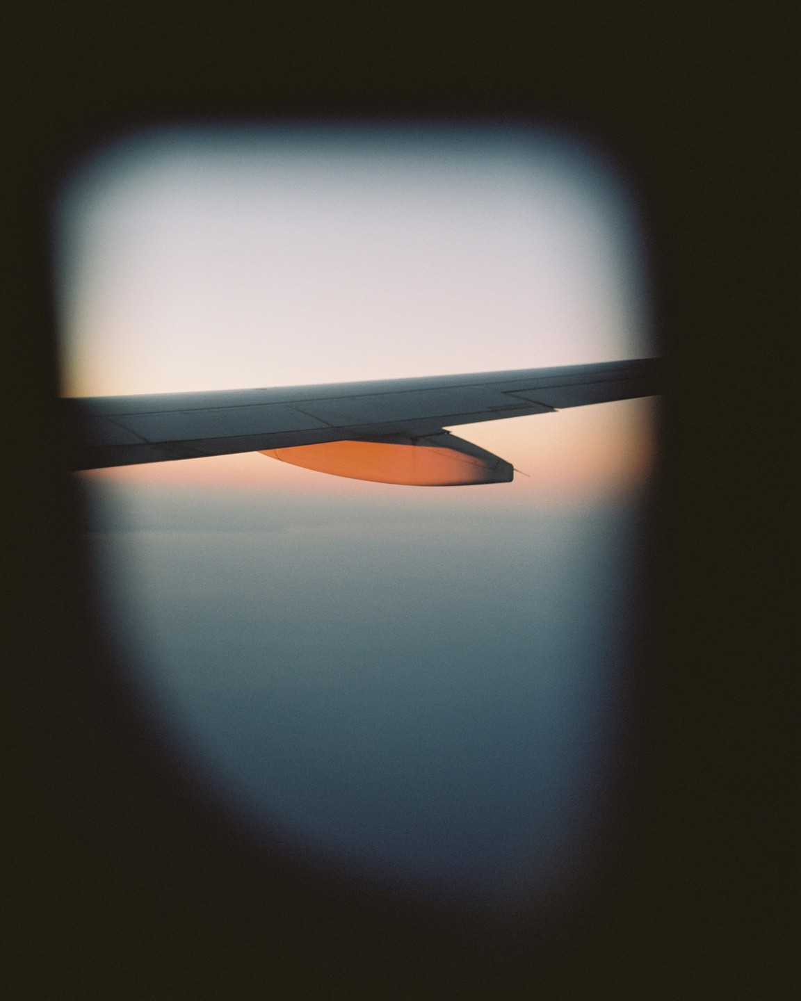 An airplane wing is visible through a window, with a sunset sky and clouds in the background.
