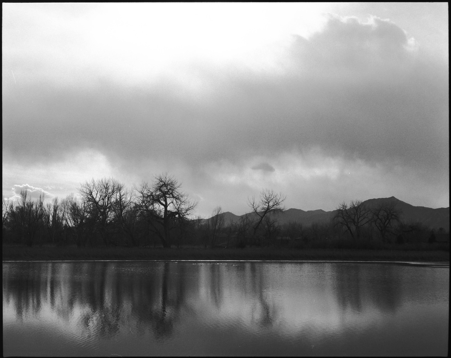 A body of water reflects a line of bare trees and mountains under a cloudy sky.