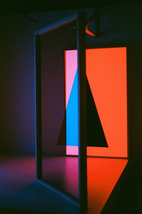A dark room is illuminated by geometric shapes of light in pink, blue, and orange, with a dark triangular shadow in the center.