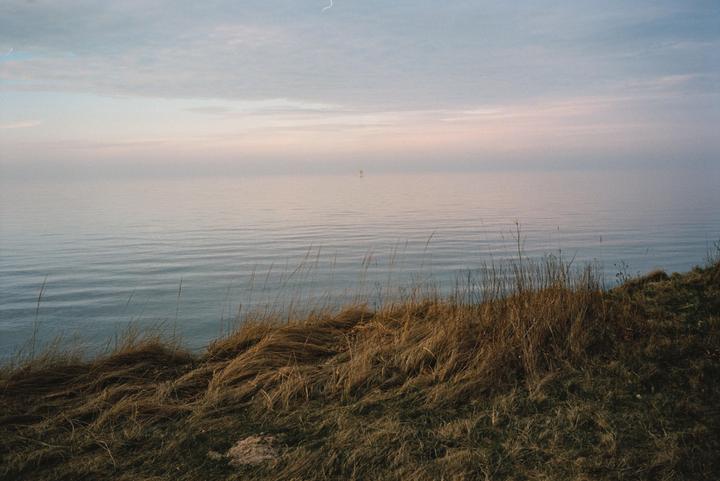 Dry grass grows on a hill overlooking a calm body of water under a hazy sky.
