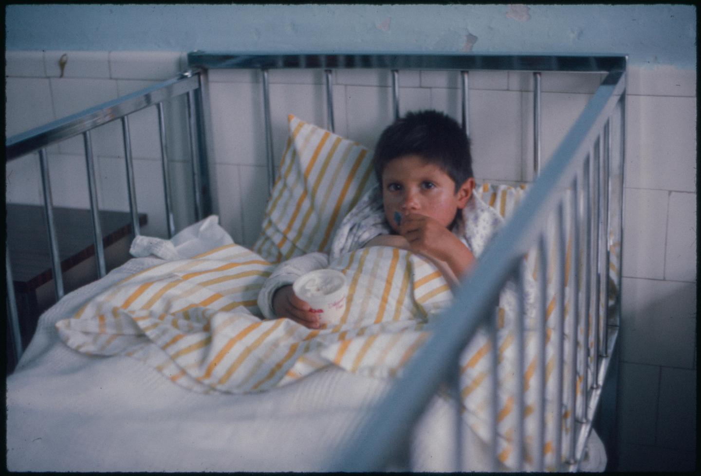 A child sits in a hospital bed with striped bedding, holding a cup and looking at the camera.