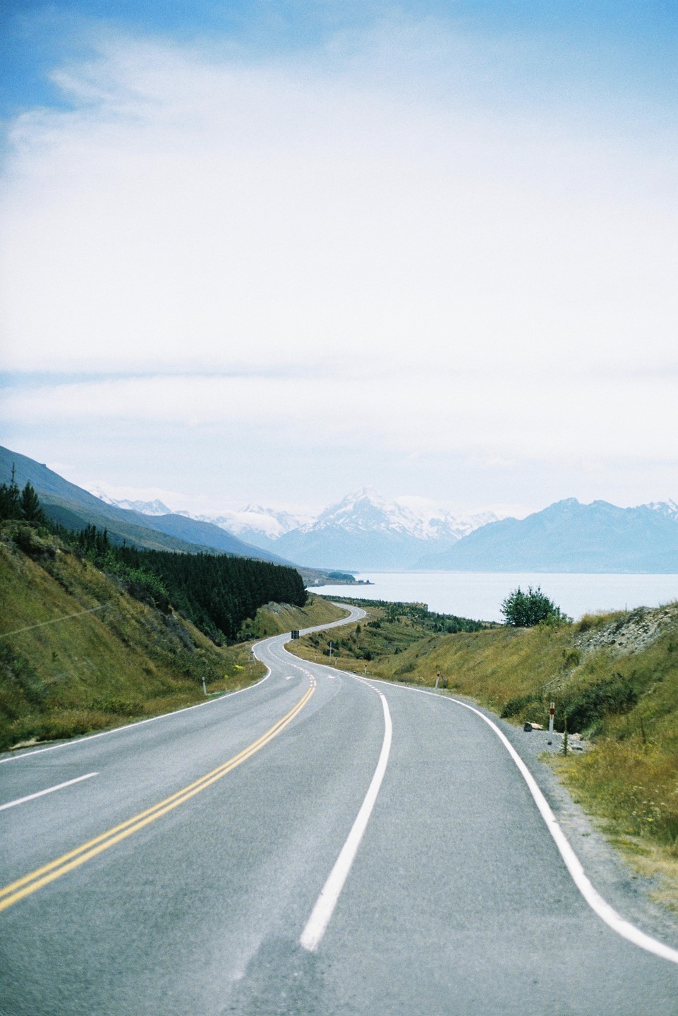 A winding road leads towards a lake and snow-capped mountains under a cloudy sky.