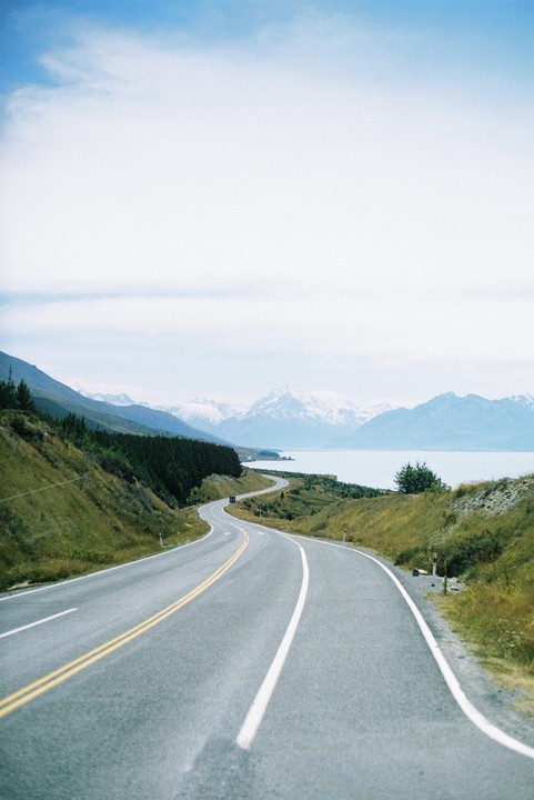 A winding road leads towards a lake and snow-capped mountains under a cloudy sky.