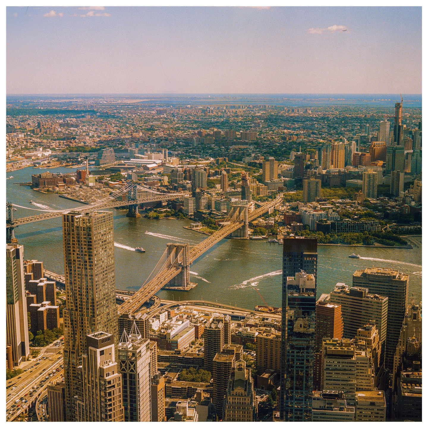 An aerial view shows the Brooklyn Bridge and Manhattan Bridge crossing a river, surrounded by a dense cityscape under a clear sky.