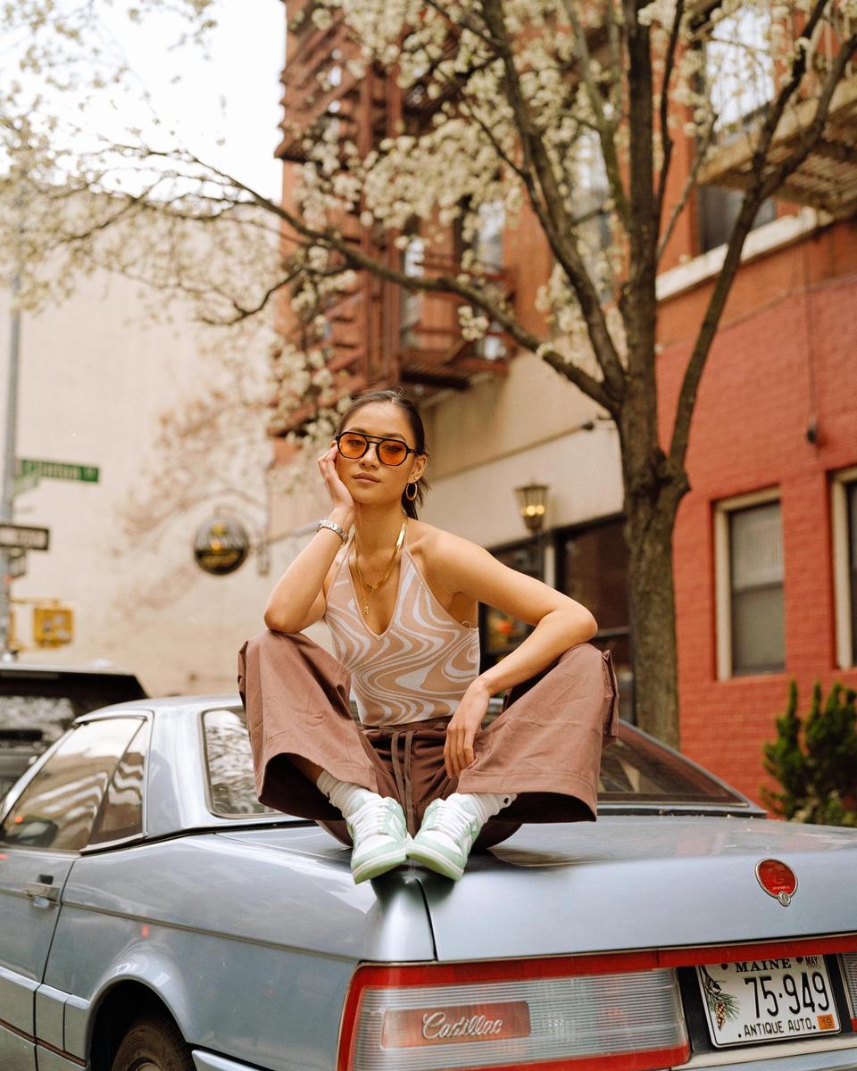 A woman wearing sunglasses and a halter top sits on the trunk of a car on a city street.