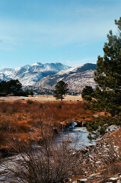 A partially frozen river flows through a field with snow-capped mountains in the background.
