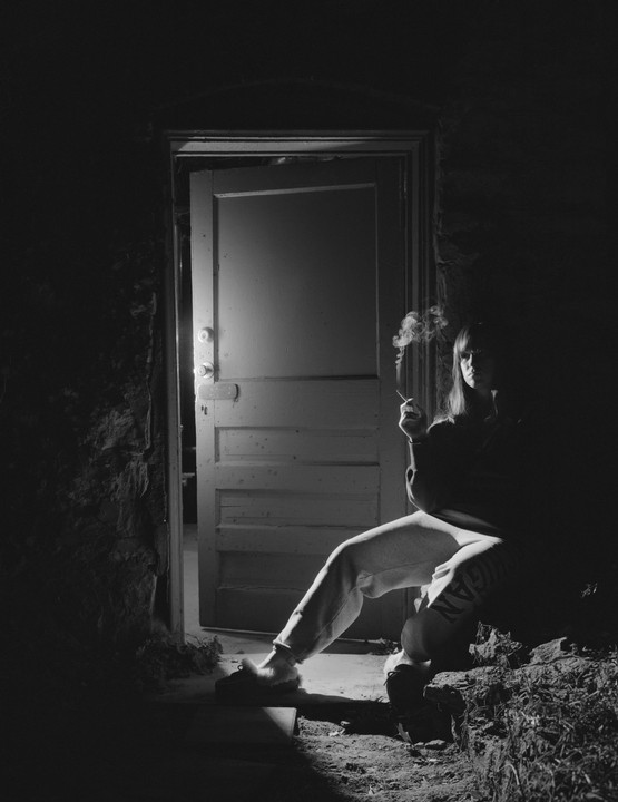 A woman sits in a doorway, smoking a cigarette, with light illuminating her from behind.