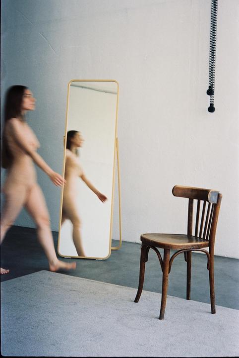 A nude woman walks past a full-length mirror, with a wooden chair in the foreground.