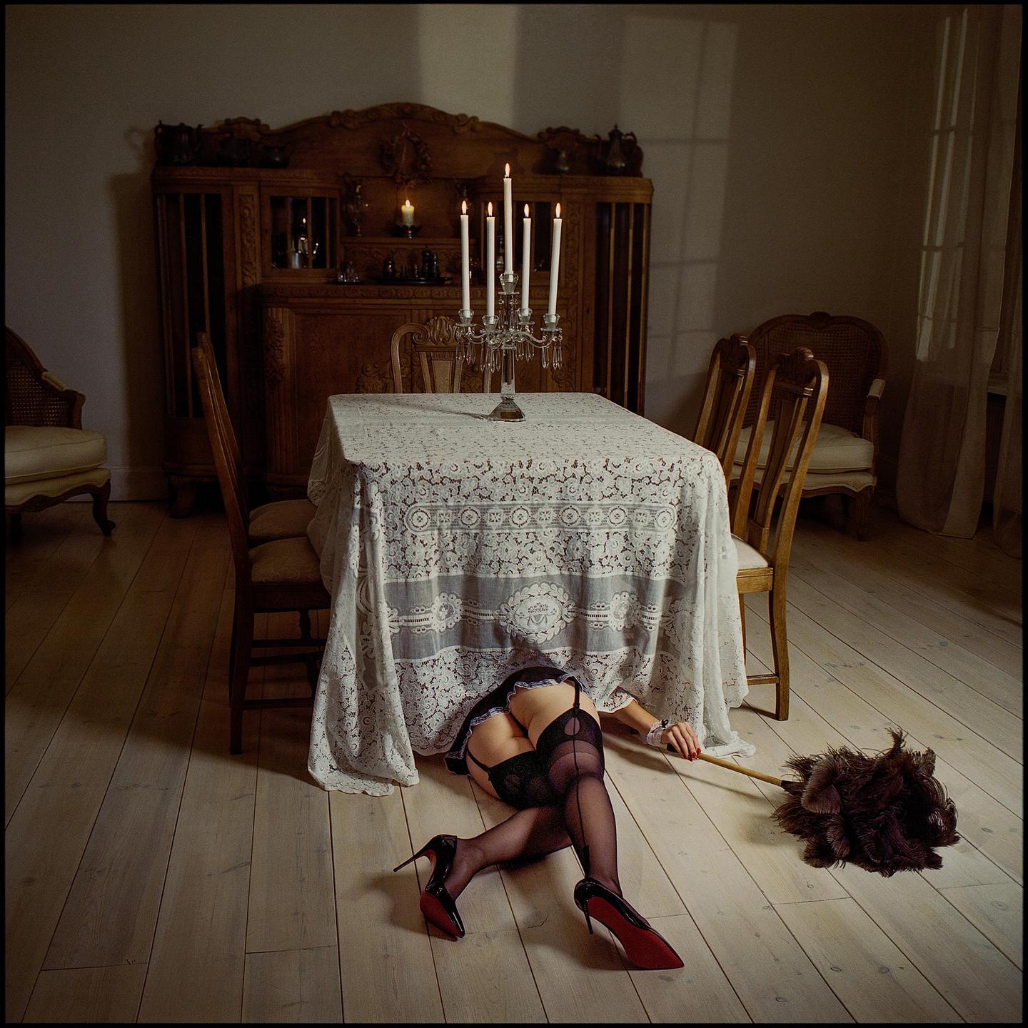 A woman's legs in stockings and high heels are visible from under a lace tablecloth, next to a feather duster on a wooden floor.