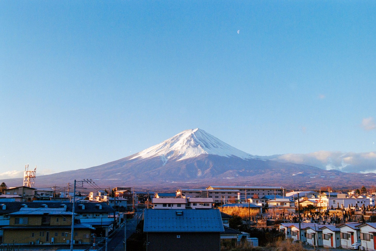 Mount Fuji rises above a town with a crescent moon visible in the clear sky.