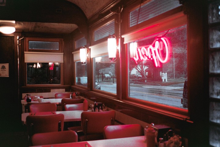 The interior of a diner with red chairs and tables is visible through a window with a neon sign.