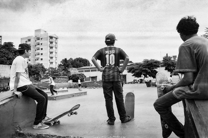 Three boys are in a skatepark, with one boy wearing a Messi jersey and two others sitting with skateboards.