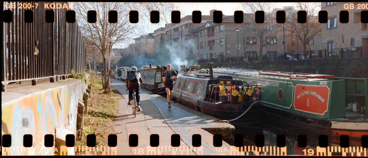 A man on a bicycle and a man running are on a path next to a canal with several boats and buildings in the background.