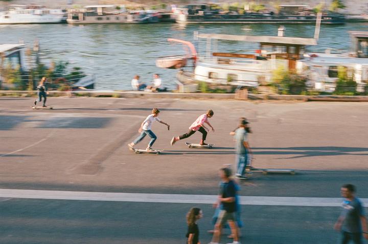 Two boys skateboard on a paved area next to a river with boats.