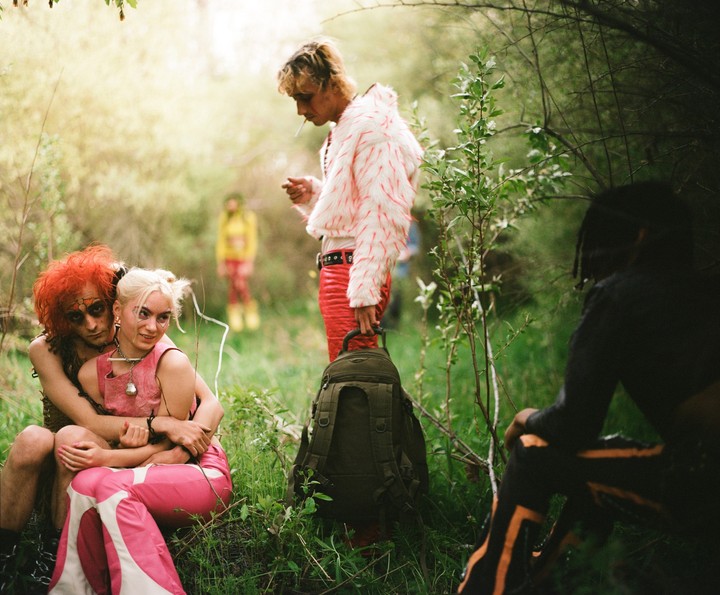 A group of people in colorful, eclectic outfits are gathered in a lush, green outdoor setting.