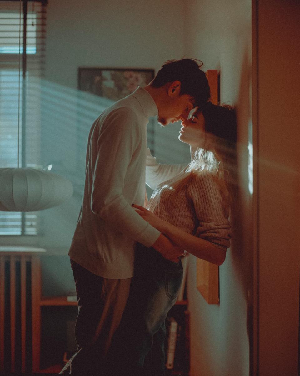 A couple stands close together, foreheads touching, with light streaming through blinds onto them.