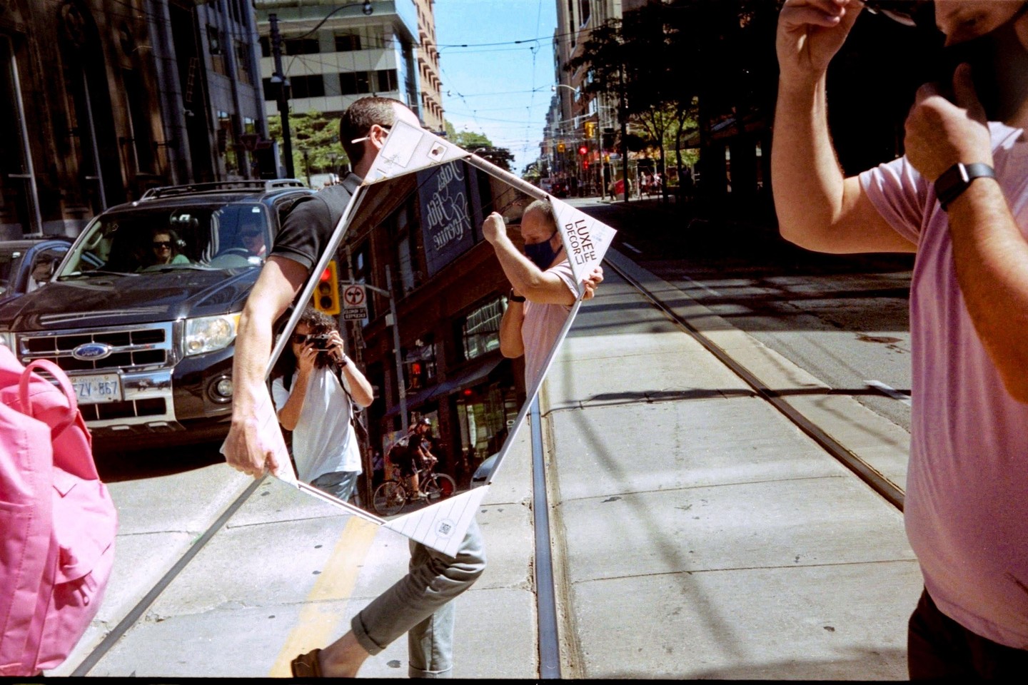 A man holds a diamond-shaped mirror reflecting a street scene with a cyclist and a person holding a camera, while another man stands to the right.