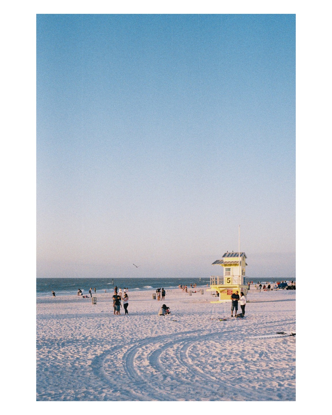 A yellow lifeguard stand stands on a white sand beach with people and the ocean in the background.