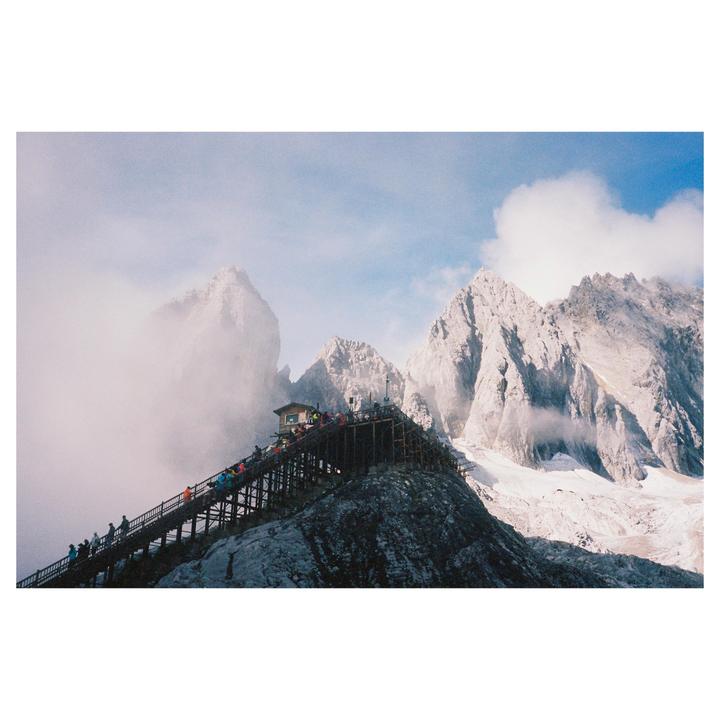 A long wooden staircase with many people leads up a mountain towards a small building, with snow-capped peaks and clouds in the background.
