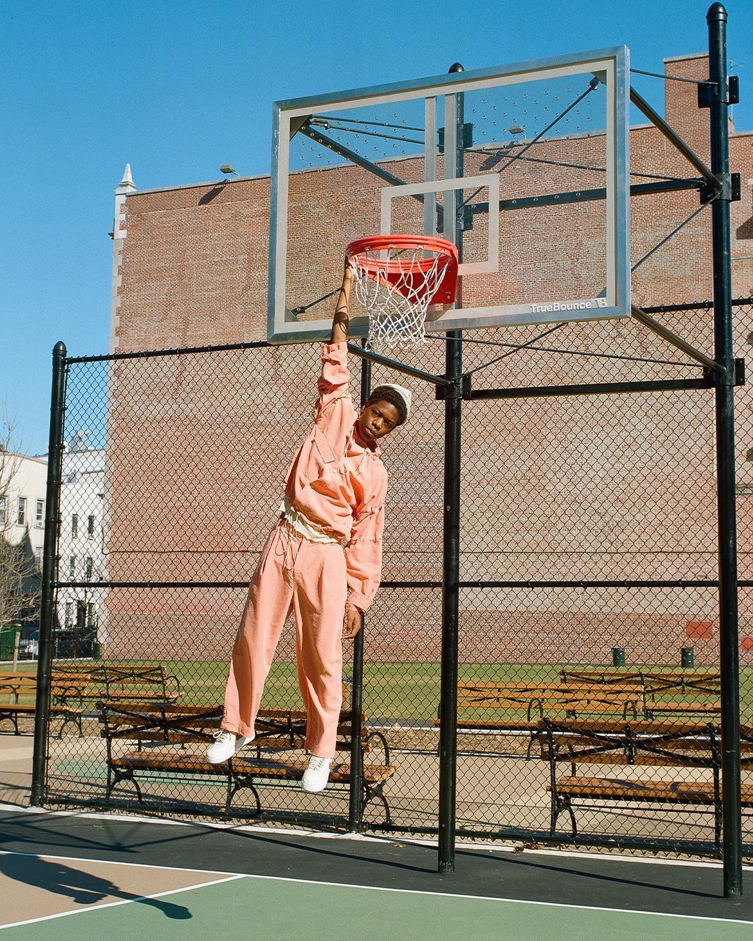 A man in a peach tracksuit hangs from a basketball hoop on an outdoor court.