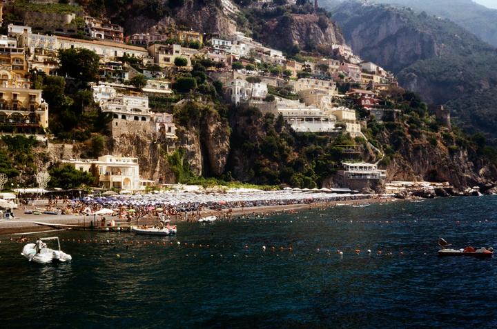 A hillside town with many buildings overlooks a crowded beach and the sea with boats.