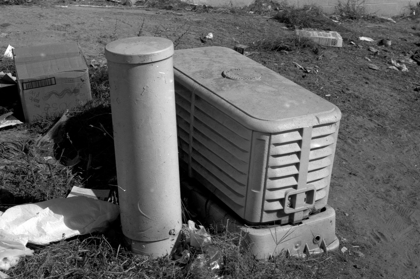 A cylindrical post stands next to a rectangular utility box on a grassy, dirt slope with scattered trash.
